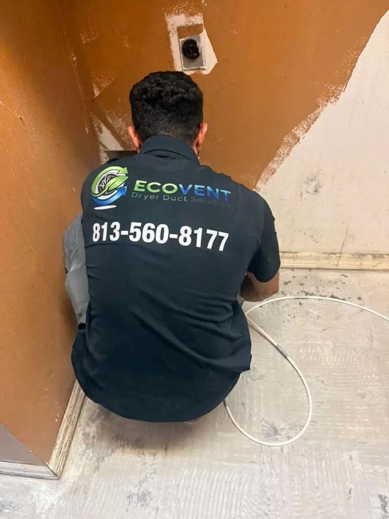 An Ecovent technician kneeling at the laundry wall where a dryer vent comes out, a white cleaning cable coiled on the floor beside him
