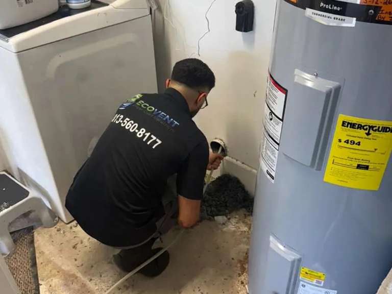 Un técnico de Ecovent agachado en un cuarto de lavado, entre la lavadora y el calentador de agua, trabajando en la conexión del ducto de la secadora en la pared, con pelusa amontonada en el piso
