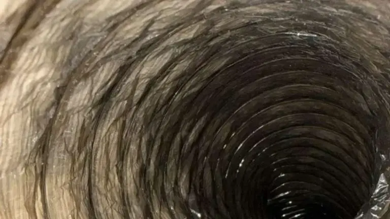 A technician in a utility room of a Florida home inserting a long flexible rotary brush into a silver dryer vent duct co